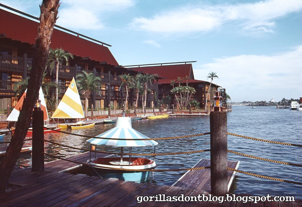 An image of a bob-a-round boat and a catamaran docked at the Polynesian Village Resort in 1971. The bob-a-round has a blue and white striped awning. You can see one of the Polynesian Resort longhouses in the background.